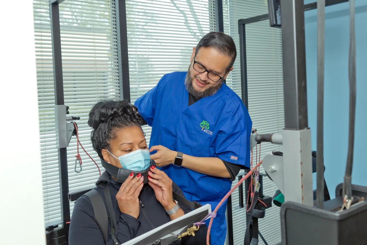 Chiropractic assistant in blue scrubs fits a neck traction harness on a masked patient seated at a machine.