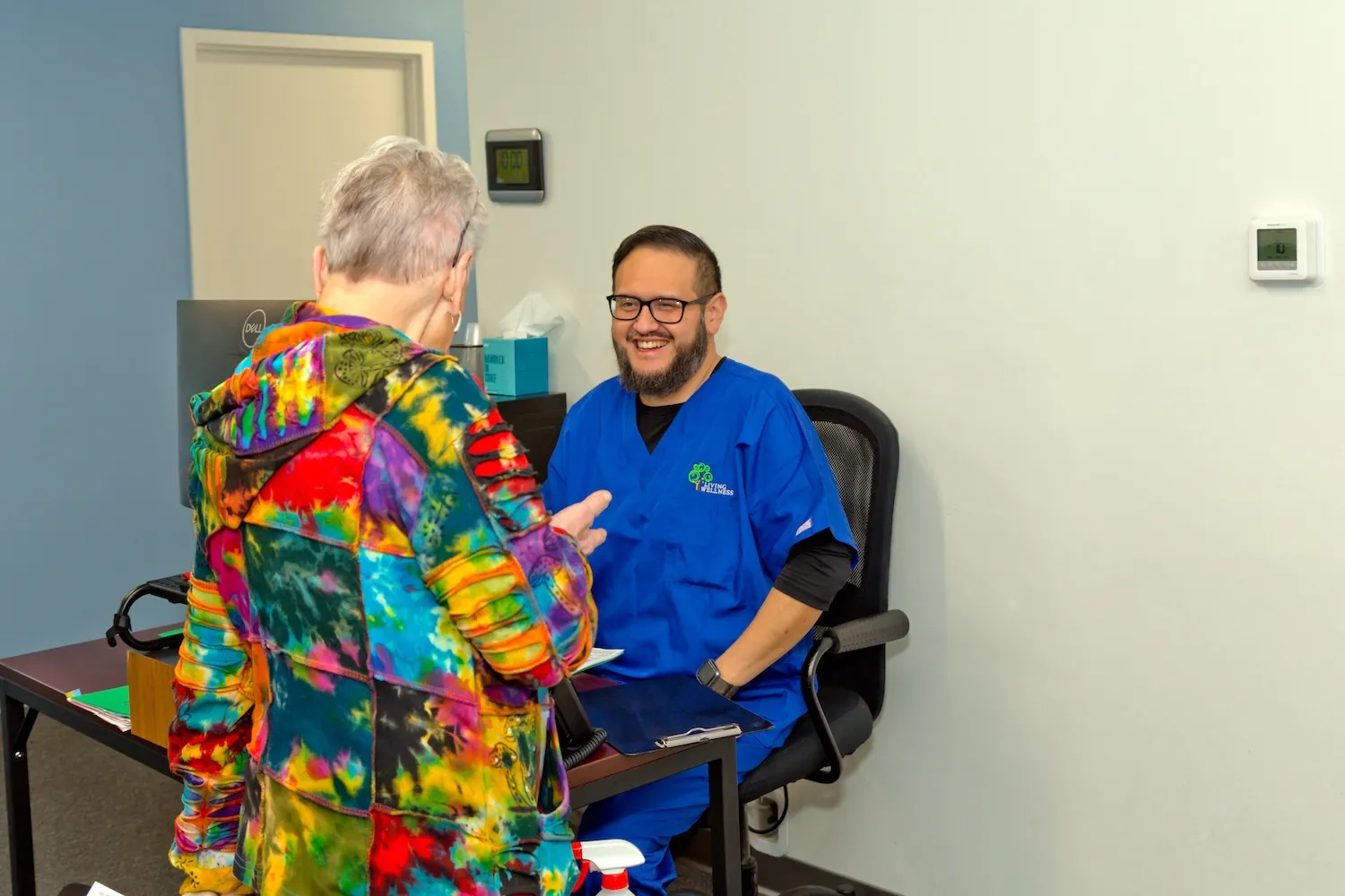 Staff member in blue scrubs smiles at a patient in a tie-dye jacket at the front desk.