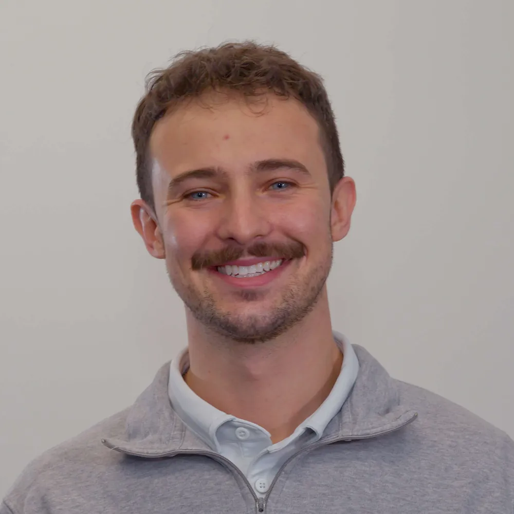 Smiling man with a mustache wearing a gray quarter-zip pullover against a plain wall.