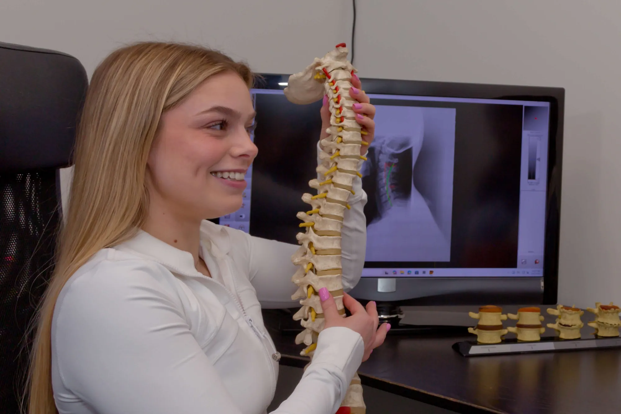 Smiling chiropractor holds a spine model beside a monitor displaying an x-ray.
