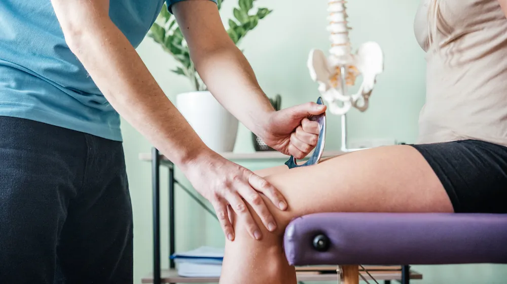 Practitioner using a handheld scraping tool on a patient's knee beside a spine model.