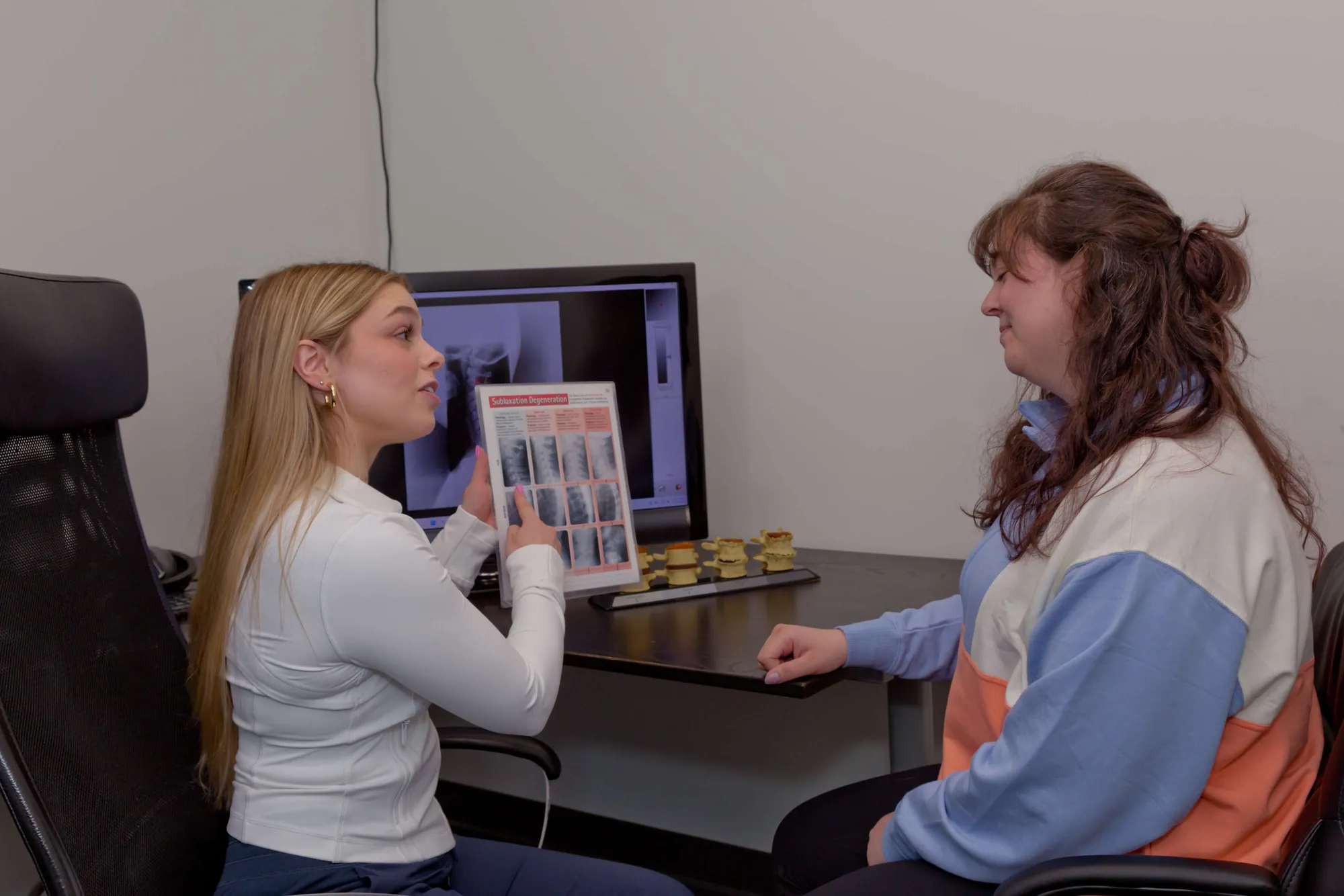 A chiropractor shows a patient a spinal degeneration card with an x-ray on the monitor behind them.