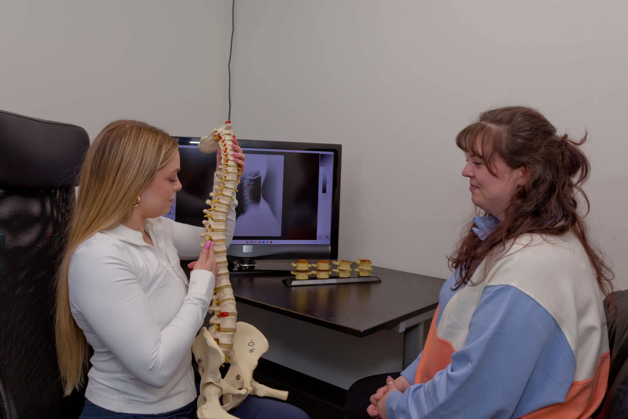 Dr. Marissa Jenney explains the spine with a model to a patient, with an x-ray on the monitor.