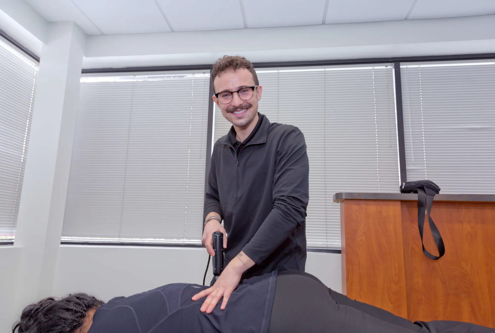 Dr. Connor Reed smiles at the camera while using a handheld adjusting instrument on a patient's back.