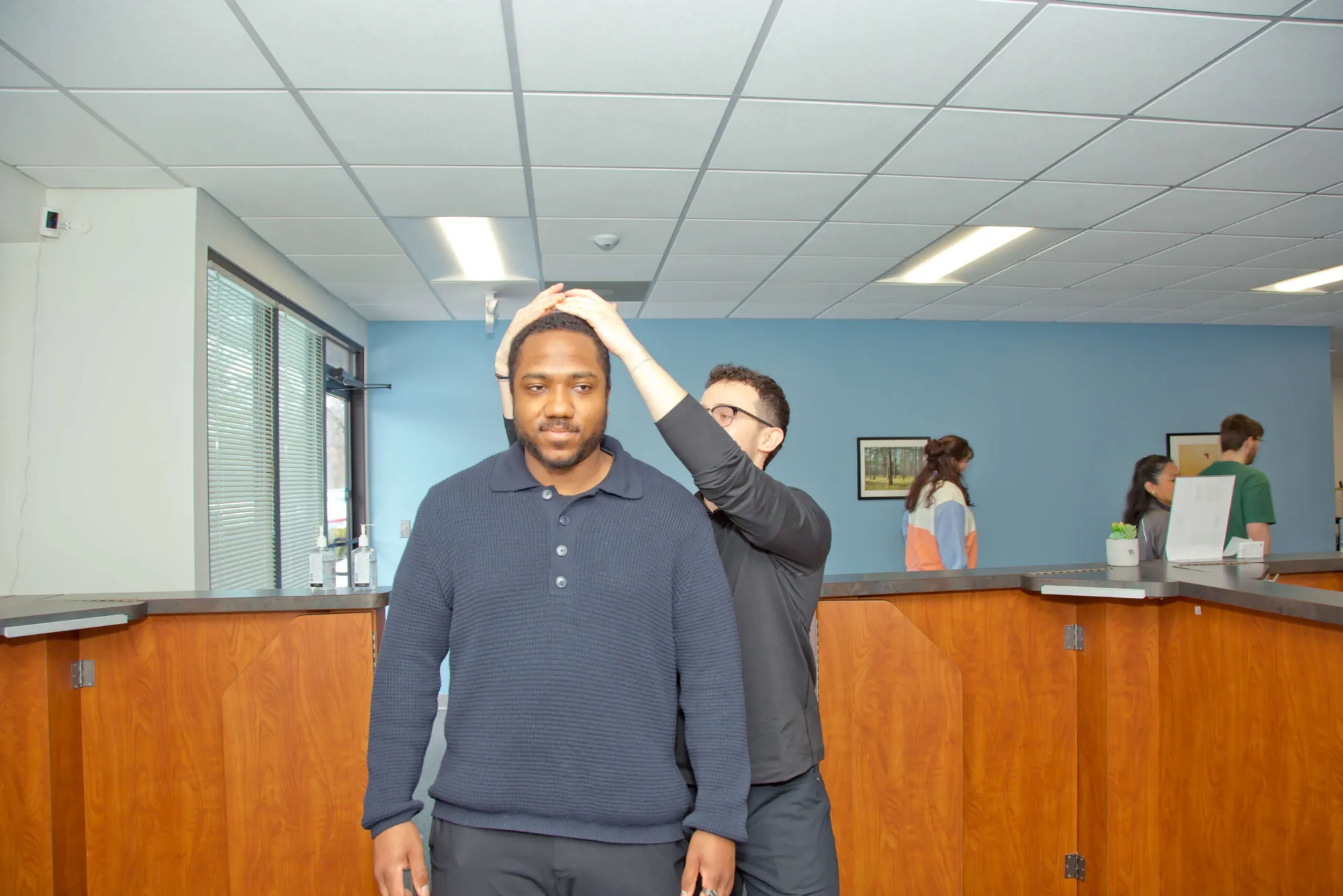 Chiropractor gently adjusts the head and neck of a standing patient as staff walk past behind them.