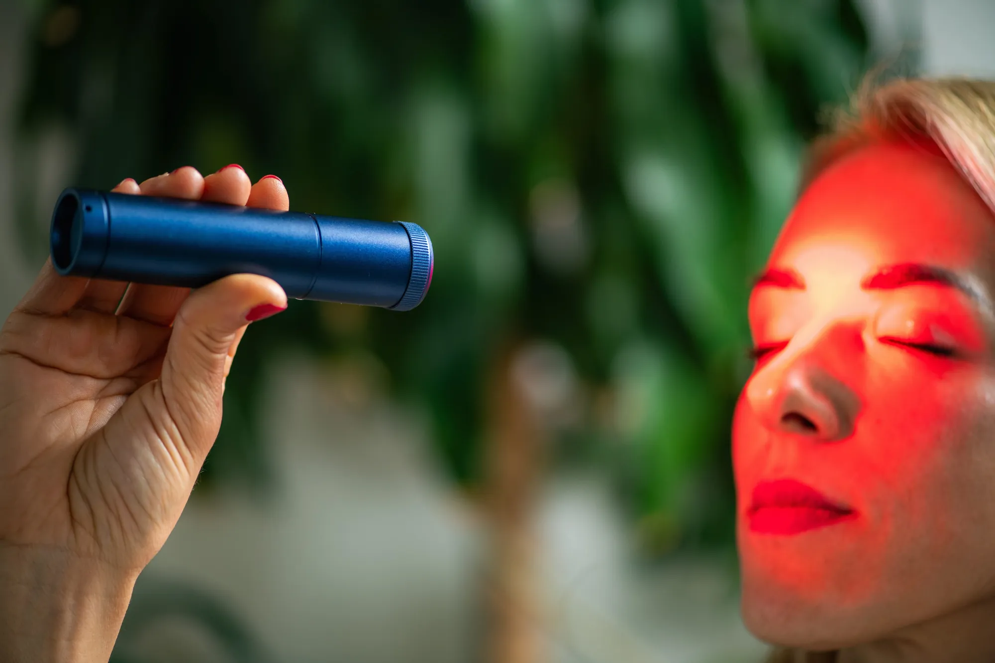 Hand holds a blue handheld red light device near a woman's face lit with red light.