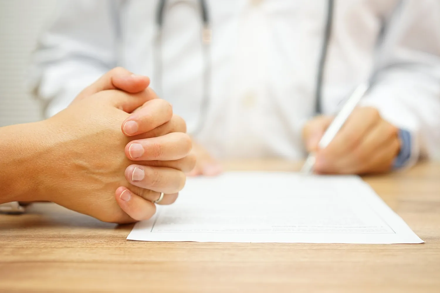 Patient's clasped hands in the foreground while a doctor in a white coat writes on a form.