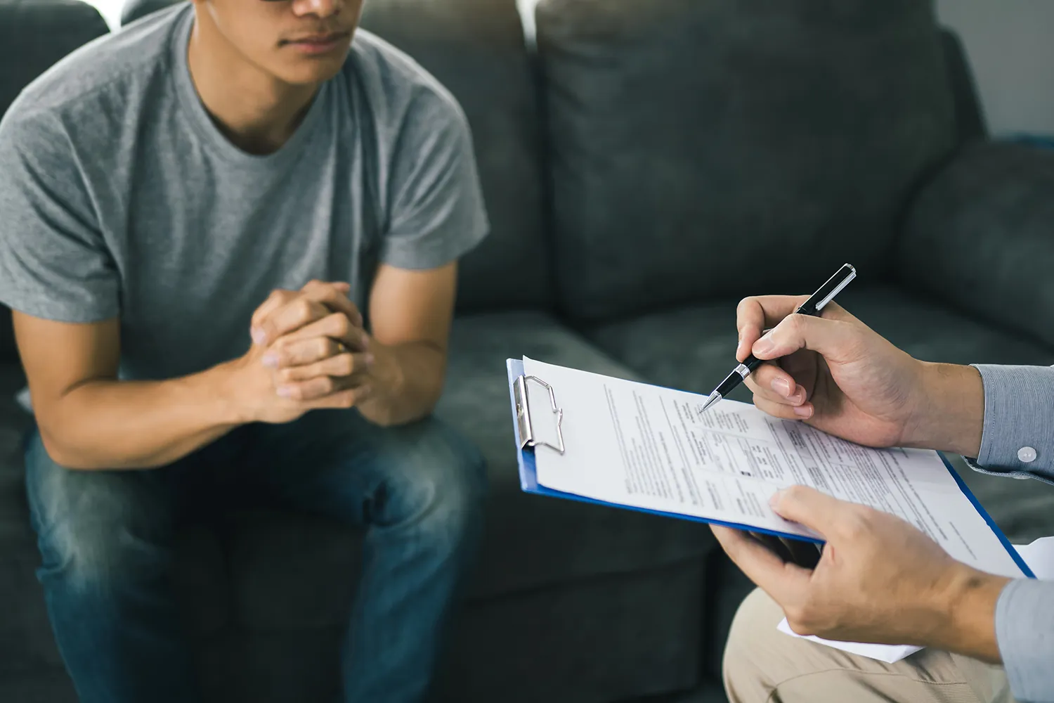 Patient sitting with clasped hands while someone fills out a form on a clipboard.