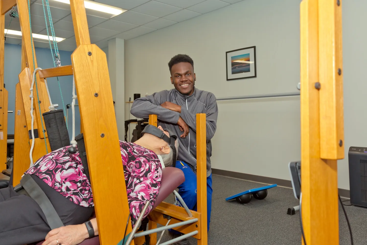 Smiling staff member leaning on a wooden traction table with a patient lying on it in the clinic.