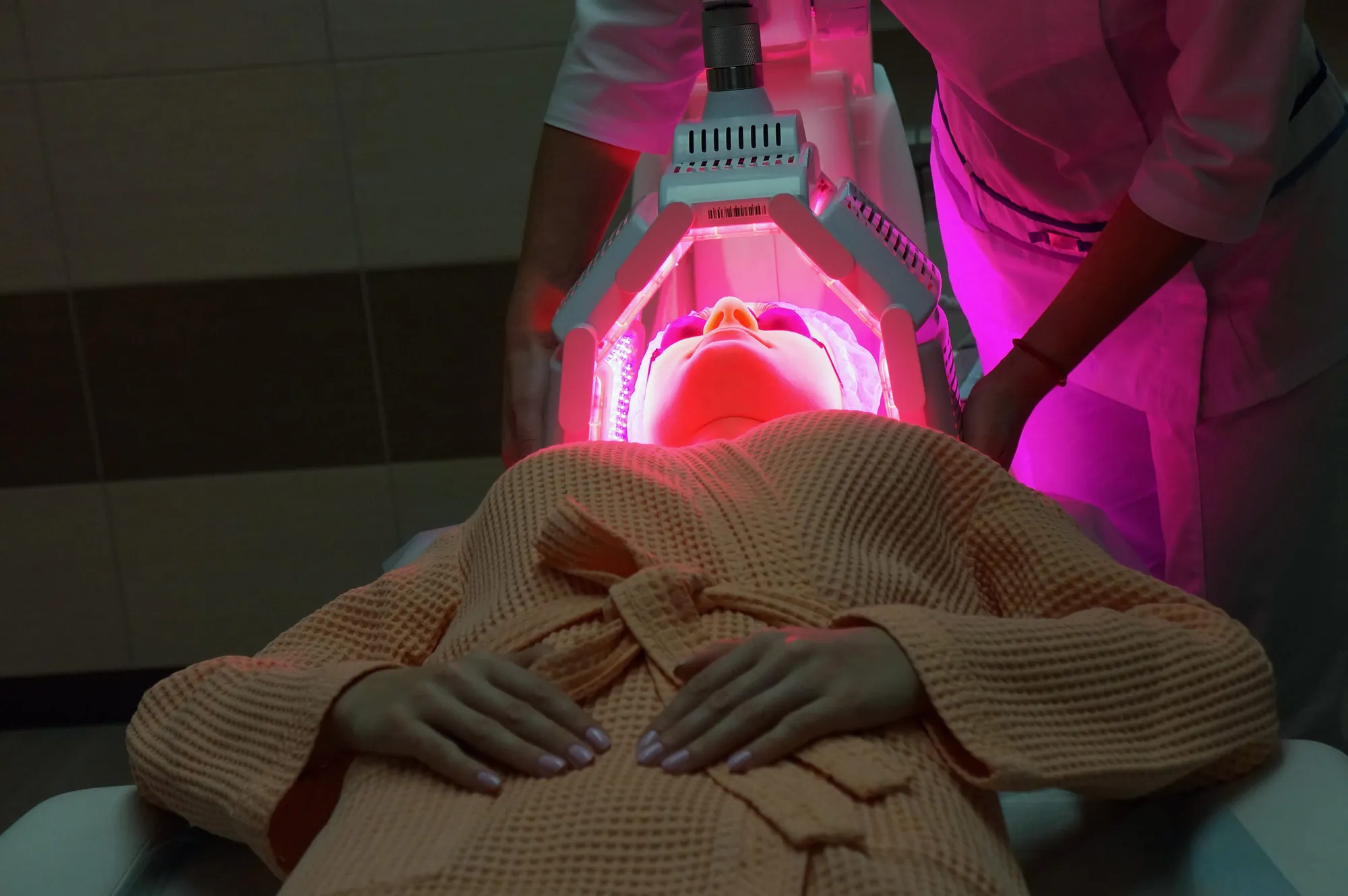 Woman lies under an overhead red light therapy device glowing over her face.