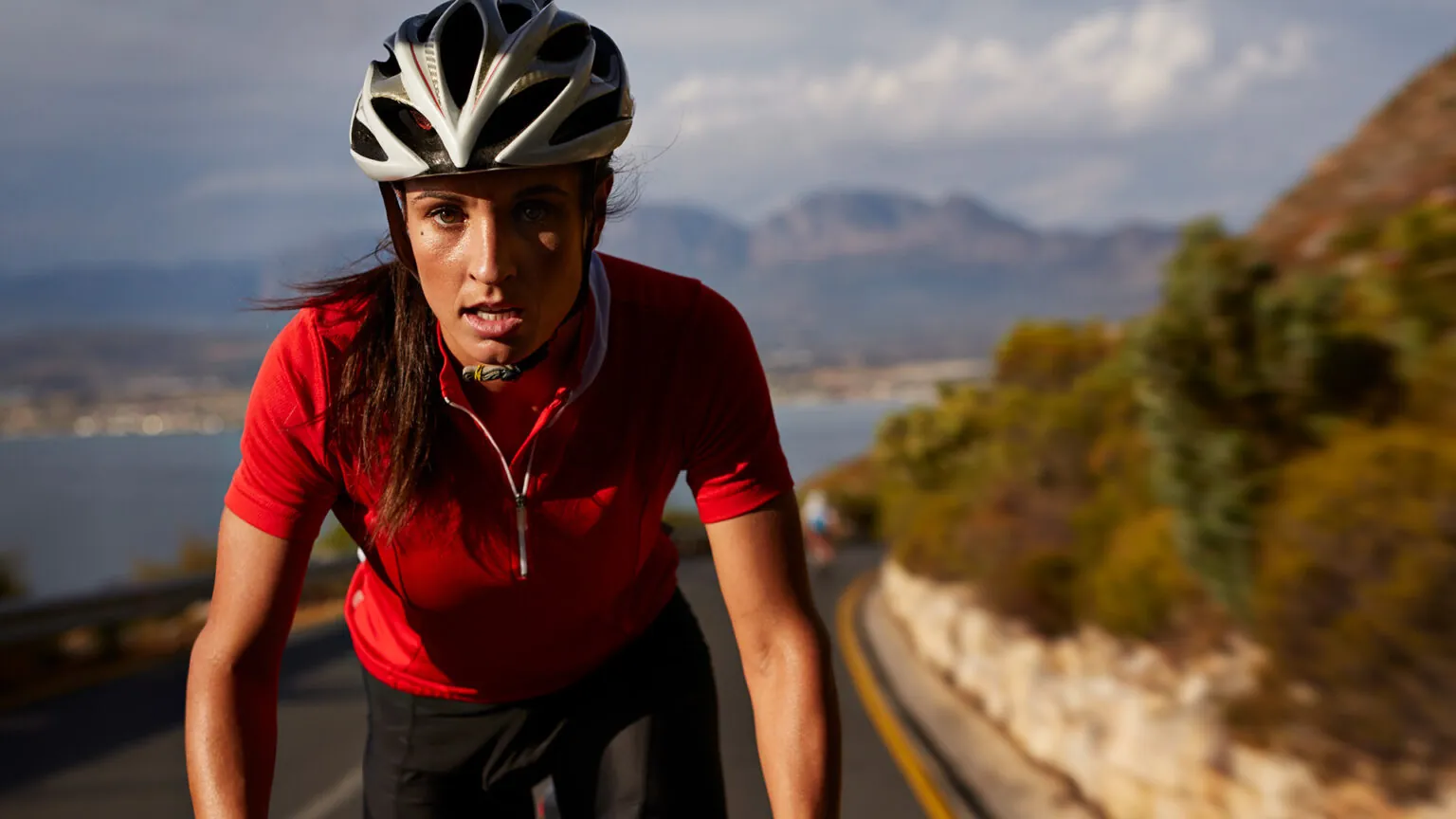 Woman in a red jersey and helmet cycling on a coastal road.