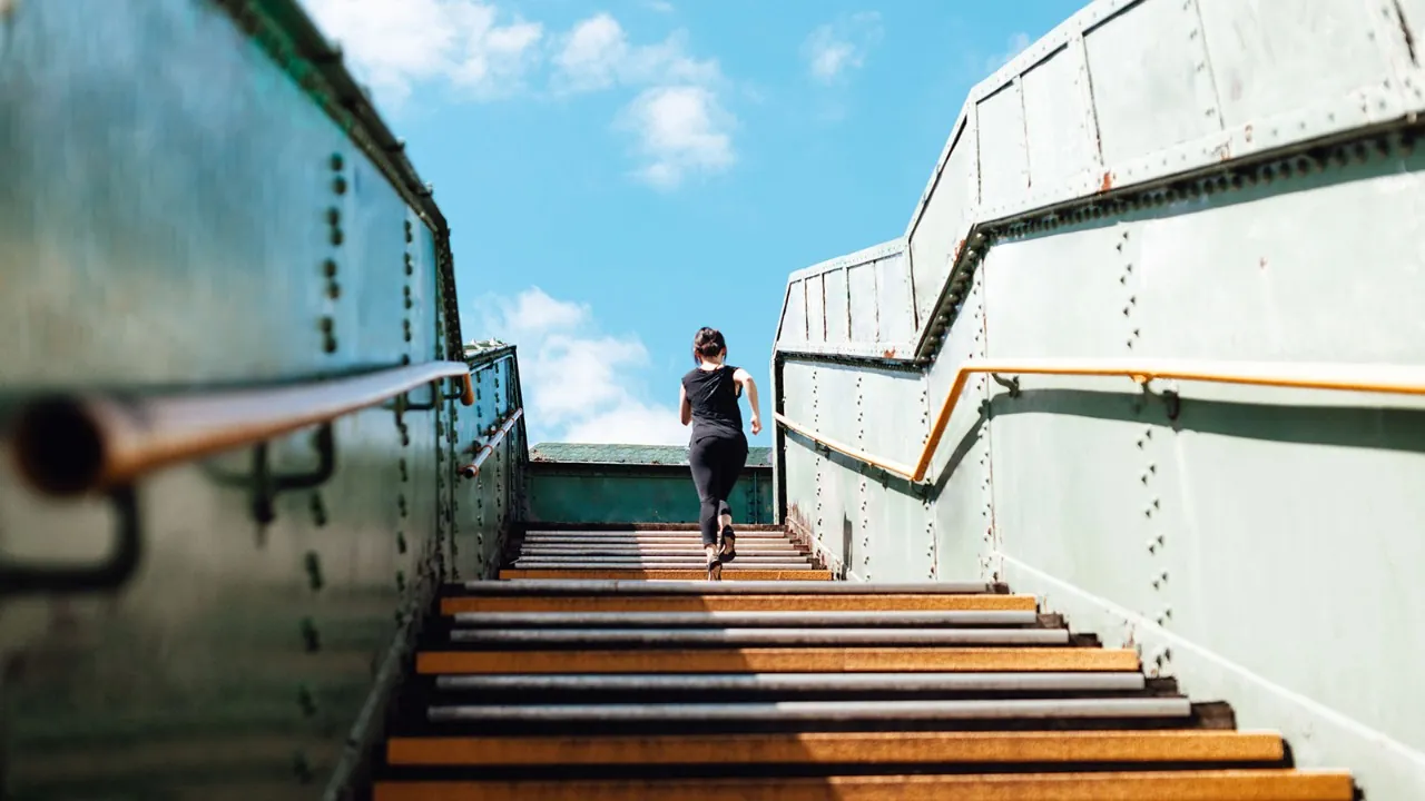Woman in black running up a flight of outdoor stairs under a blue sky.
